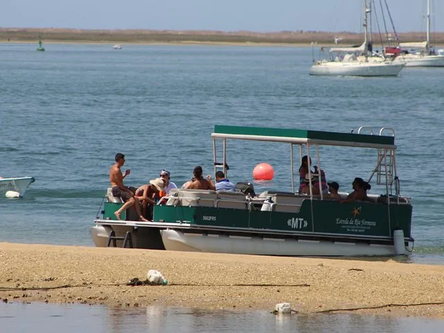 Barco turístico en una playa de arena con pasajeros y veleros al fondo